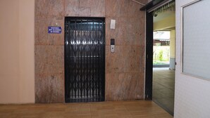 Ascensore con un cartello che indica che non sono ammessi visitatori nelle stanze, una tastiera numerica e una porta che dà su un corridoio con vista su aree verdi.