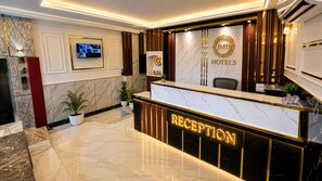 A hotel reception area with a marble floor, a reception desk, a television mounted on the wall, and a logo with 'JMD HOTELS' on the wall. - Hotel Paradise International by JMD Group (New Delhi)