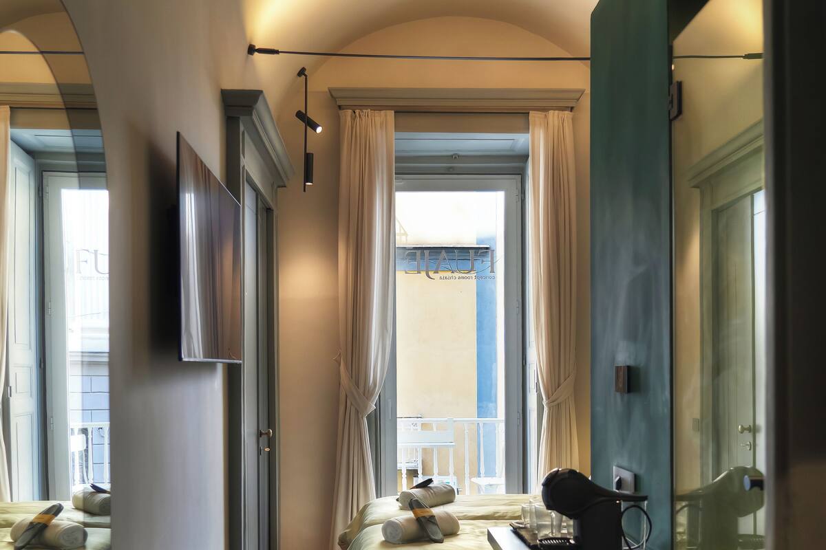 A hotel room with a bed, a desk with a computer, a chair, a TV mounted on the wall, and a window with curtains.