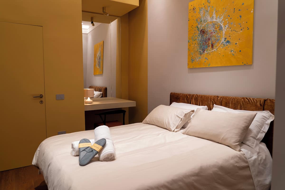 A hotel room with a bed, a desk, and a painting on the wall.