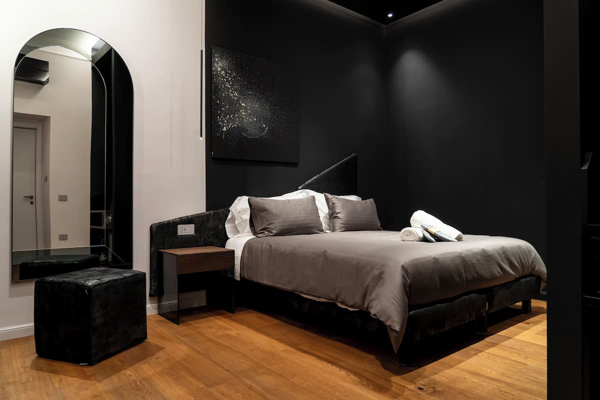 A modern bedroom with a bed, a nightstand, a black ottoman, a large mirror, and a wall art piece.