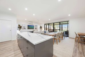 A modern kitchen with a marble island, dining area with wooden furniture, and large windows offering a view.
