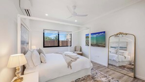 Une chambre à coucher comprenant un lit, une lampe de chevet, un ventilateur de plafond, une fenêtre et un grand miroir.