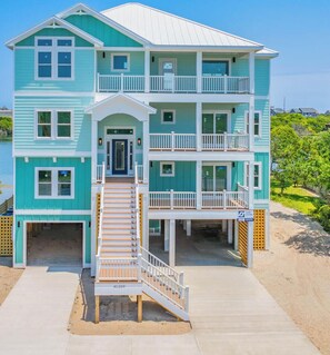 A multi-story beachfront house with a front porch, a staircase, and a visible garage.
