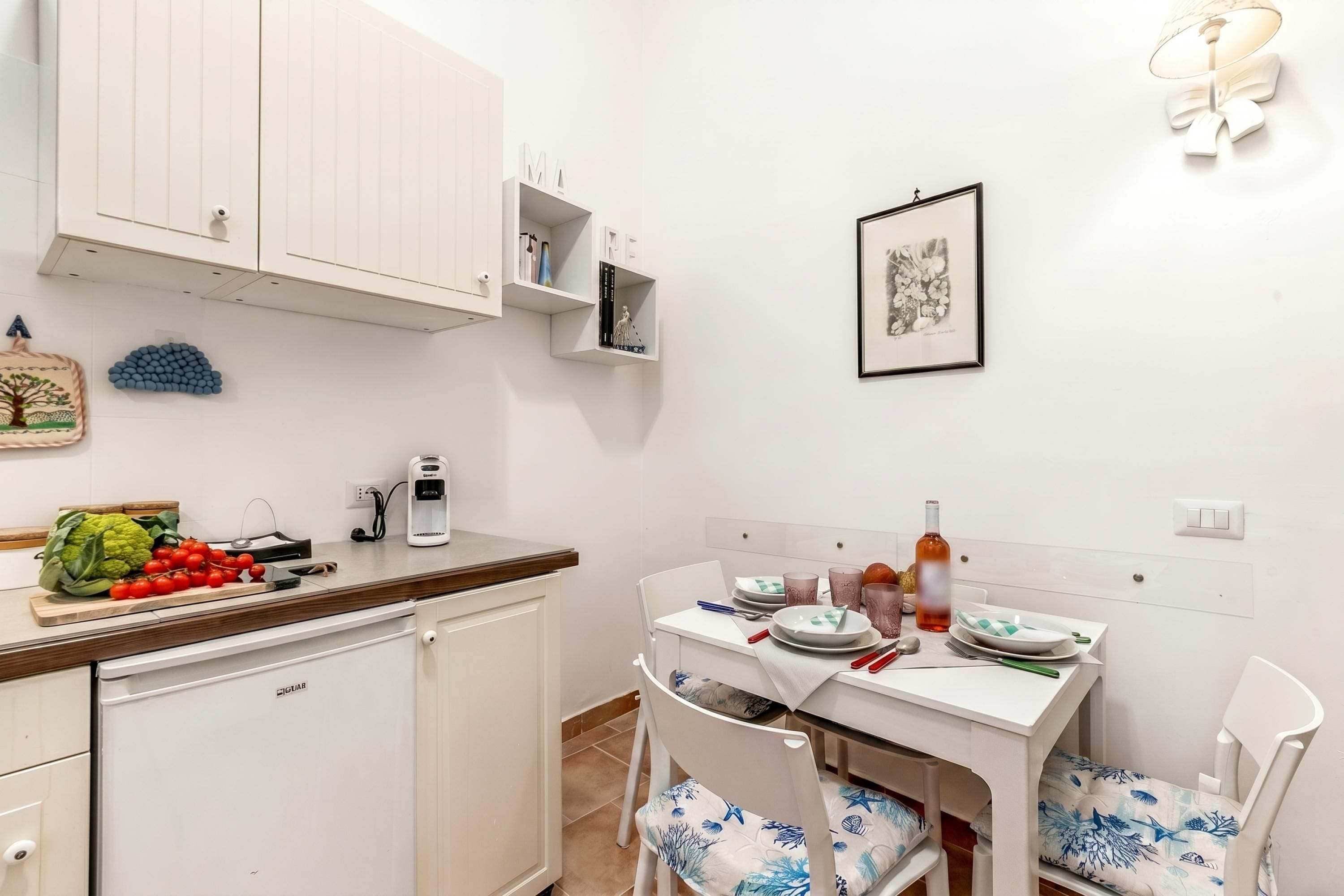 Een kleine keuken met witte kasten, een eettafel met stoelen en een kleine koelkast.
