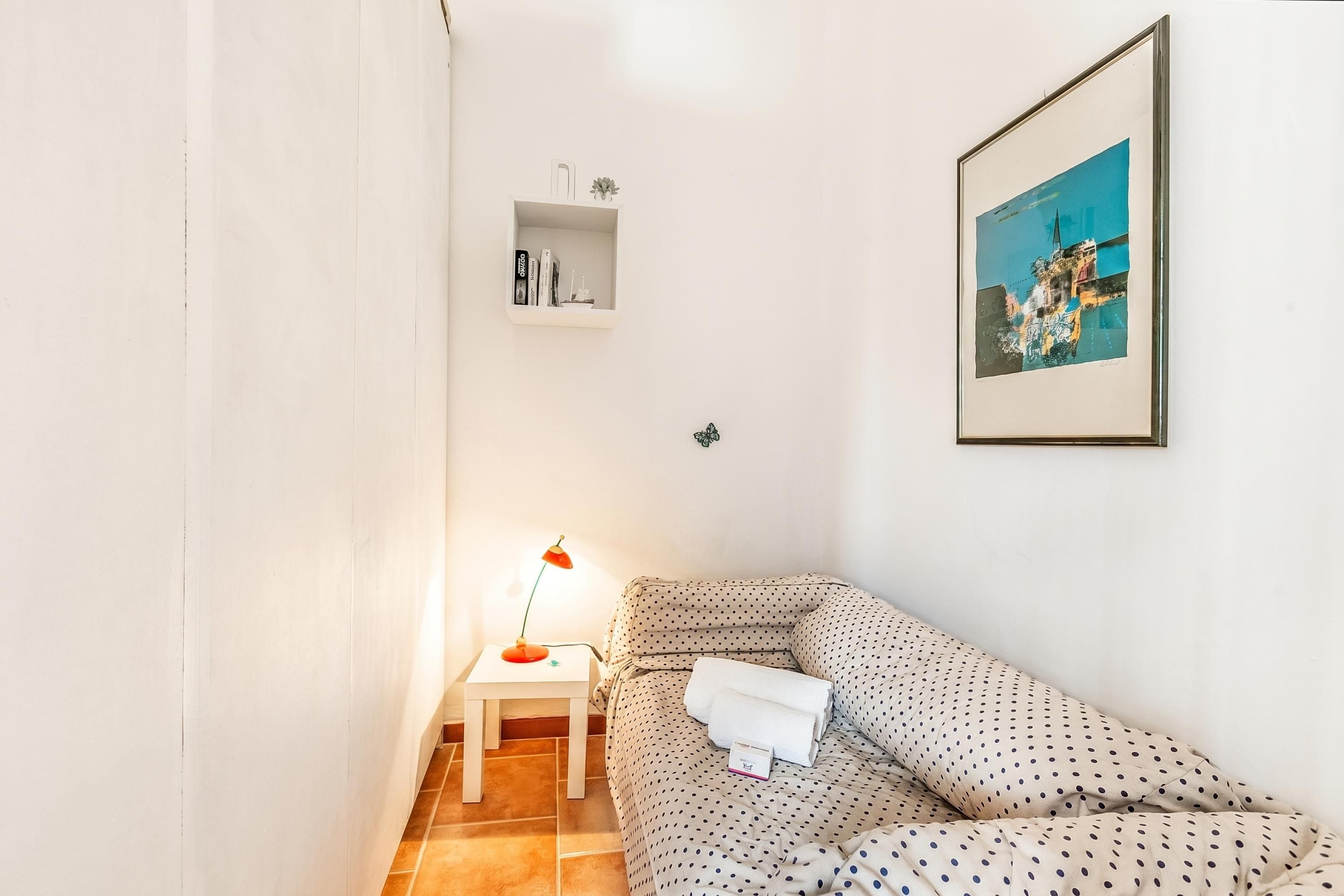 Een kleine, netjes ingerichte kamer met een bed, een nachtkastje met een lamp, een lijst met een foto aan de muur, en een plank met boeken en een kleine plant.