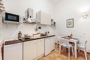 Egy praktikus, kis méretű konyha, amelyben mikrohullámú sütő, tűzhely és fehér szekrények találhatók. Van egy étkezőasztal székekkel és egy kis hűtőszekrény is.