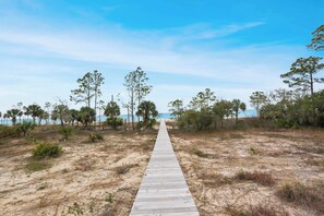 Ein hölzerner Steg führt zu einem Strand mit wenig Vegetation und blauem Himmel.