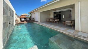 Une piscine avec un espace patio, une table à manger et une cuisine visible à travers une porte ouverte.