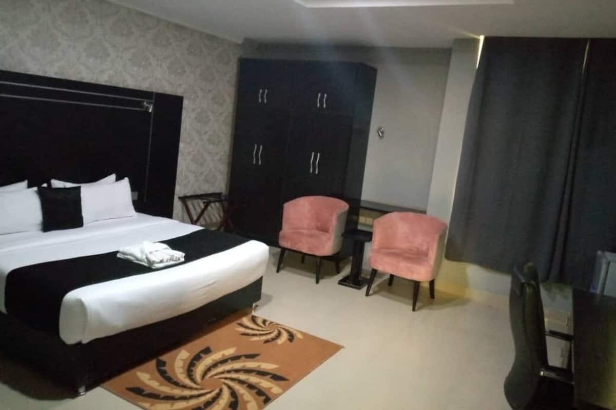 A hotel room with a large bed, two pink chairs, a dark wardrobe, and a small table.