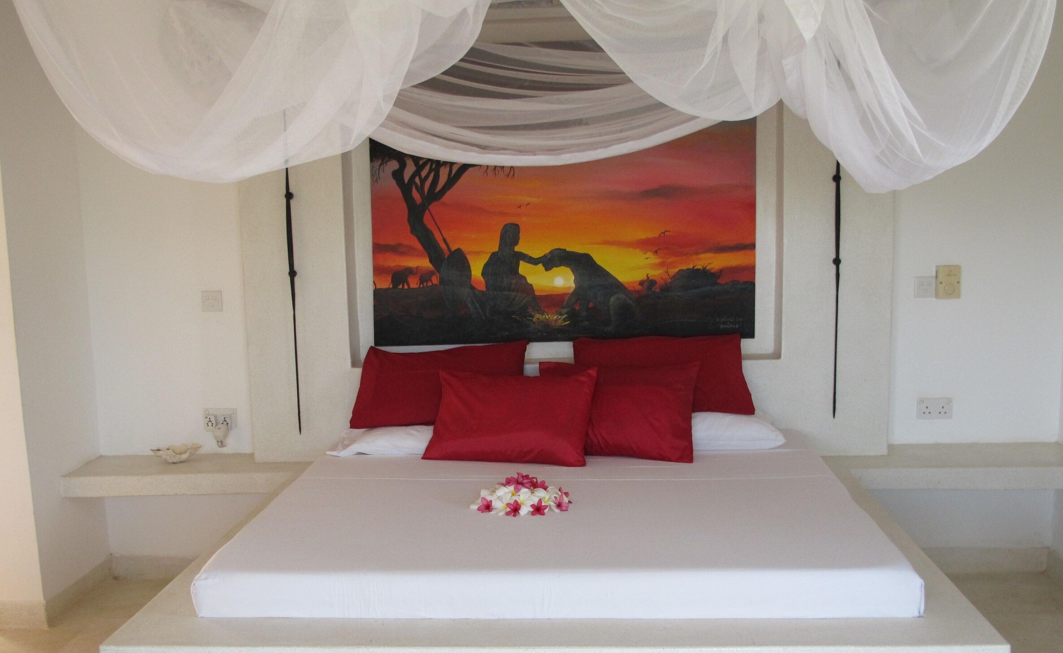 Een slaapkamer met een bed, rode kussens, een wit baldakijn en een schilderij van een zonsondergang aan de muur.