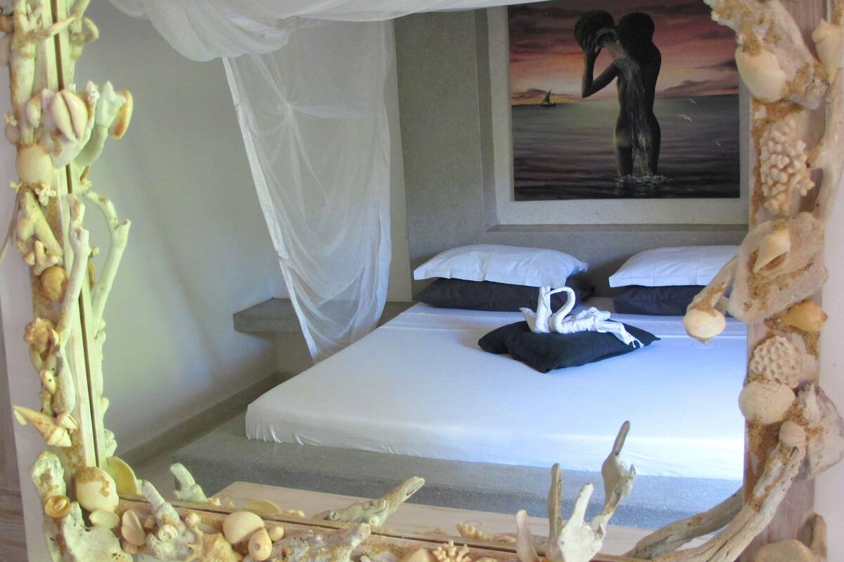 A bedroom with a bed, white bedding, a painting of two people on the wall, and a canopy over the bed.
