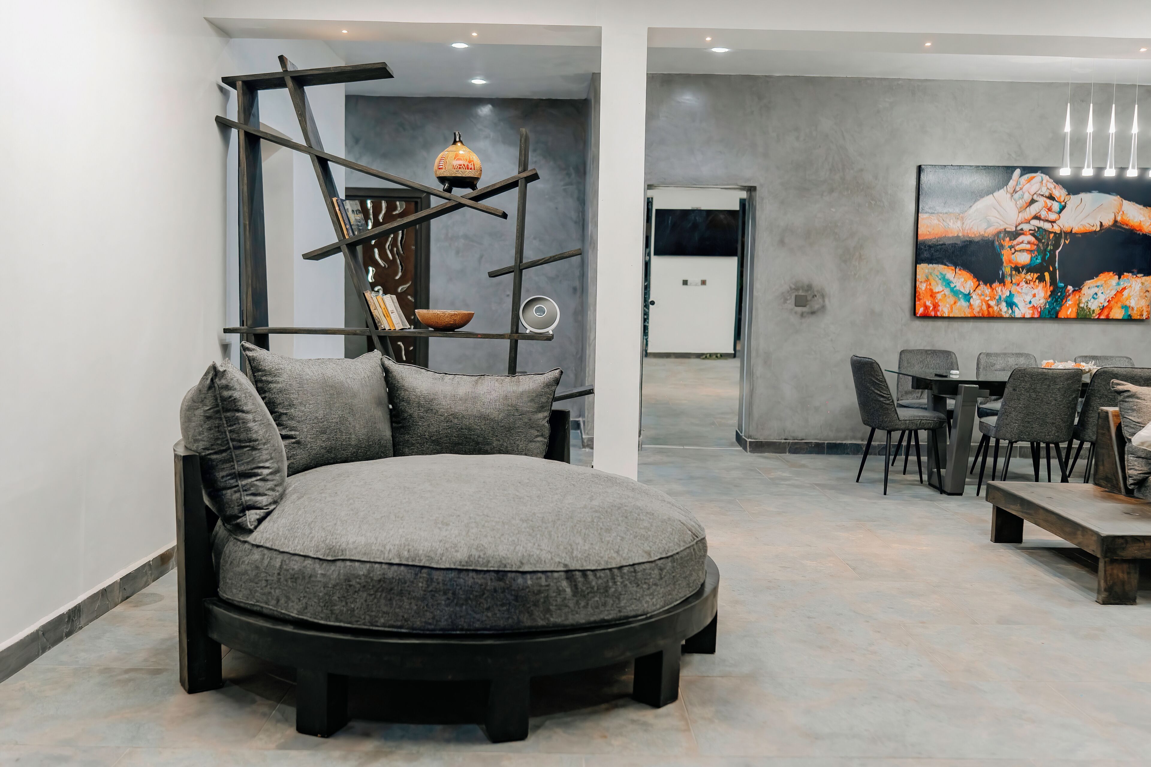 A modern living room with a round grey sofa, a wooden ladder shelf with decorative items, a dining area with grey chairs, and a large abstract painting on the wall.