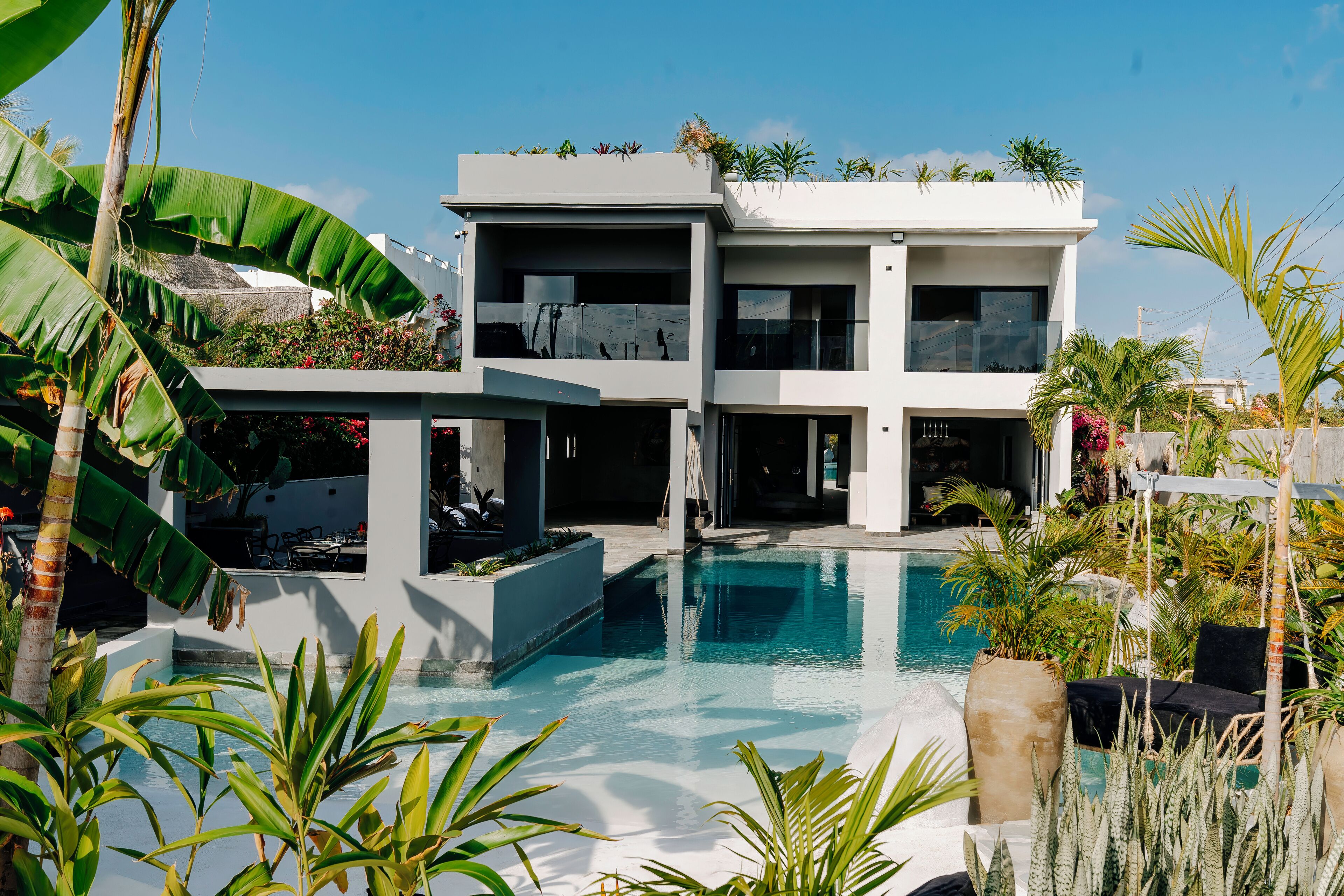 A modern two-story house with a swimming pool, surrounded by tropical plants and palm trees.