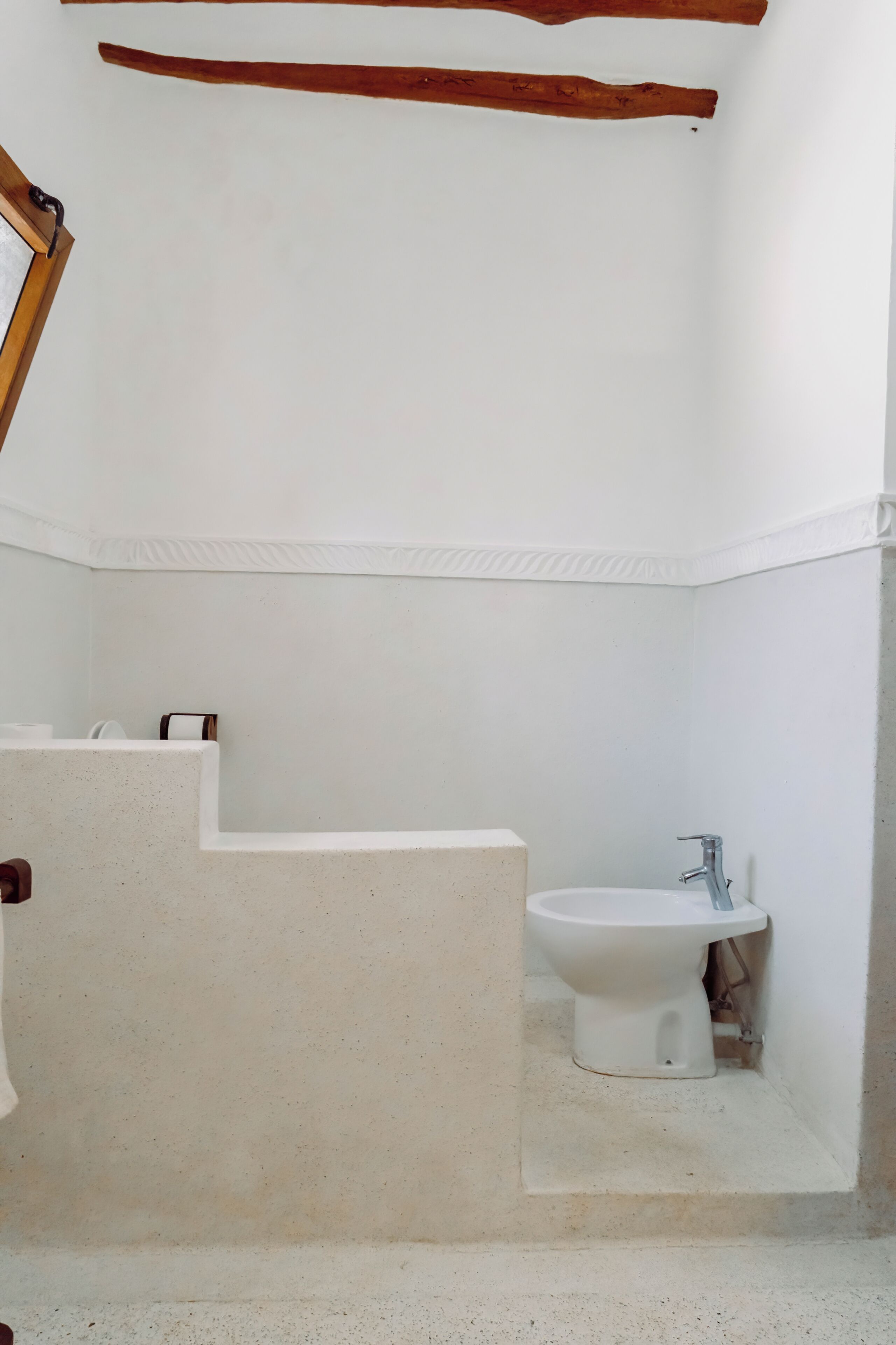 A small, white bathroom with a toilet and bidet, a wooden beam above the toilet, and a white wall with a textured pattern near the floor.