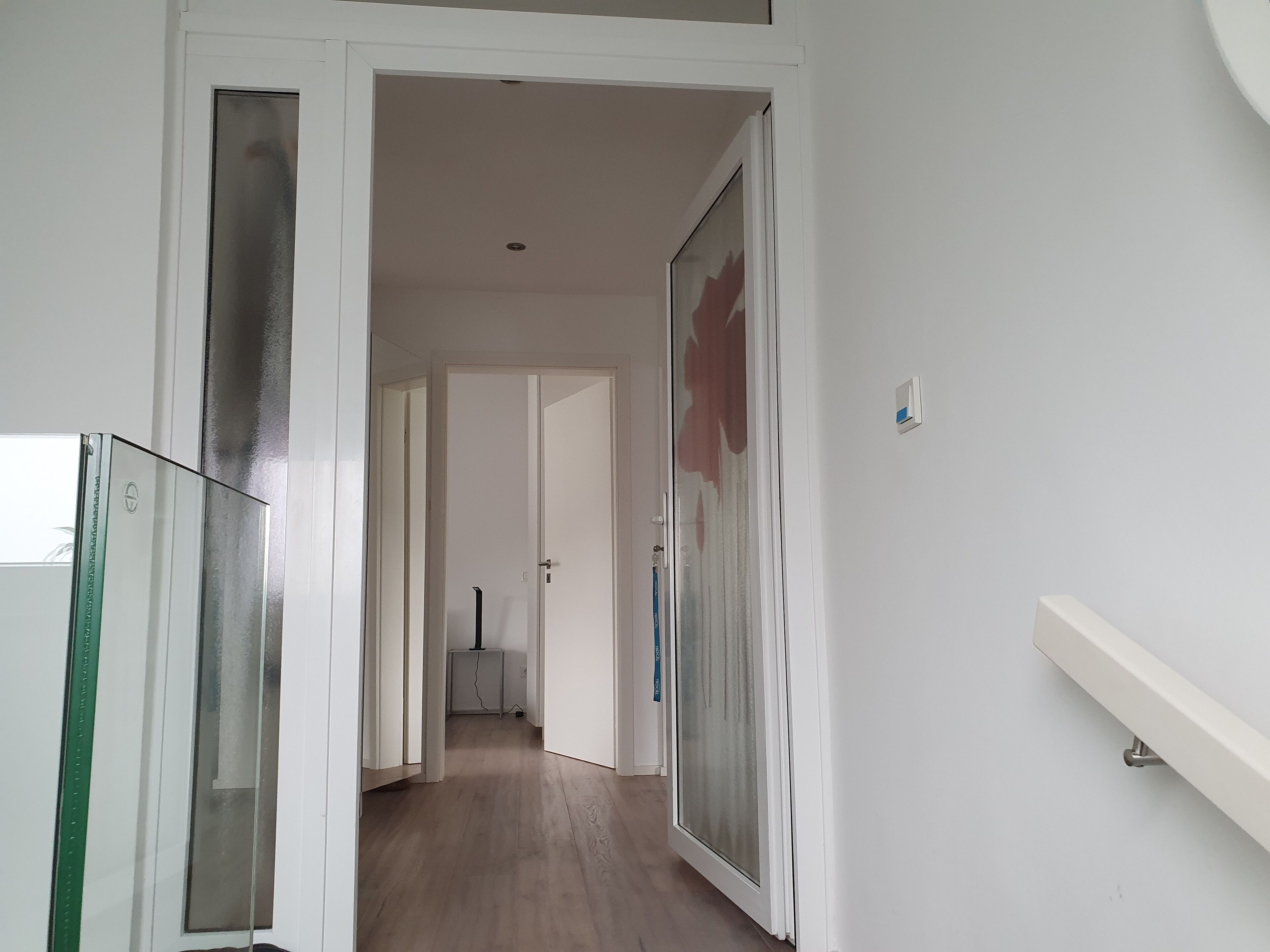 Un pasillo con una puerta de vidrio a la izquierda, una puerta blanca en el centro y una pared blanca a la derecha.