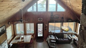 Interior de una cabaña con techo alto, paredes de madera, zona de comedor, sala de estar y grandes ventanales con vistas al exterior.