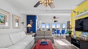 Ein modernes Wohnzimmer mit einem weißen Sofa, einem roten Couchtisch und einem gestreiften Sofa. Zwei Deckenventilatoren, ein Fernseher und ein großes Fenster mit Blick auf die Stadt sind ebenfalls vorhanden.