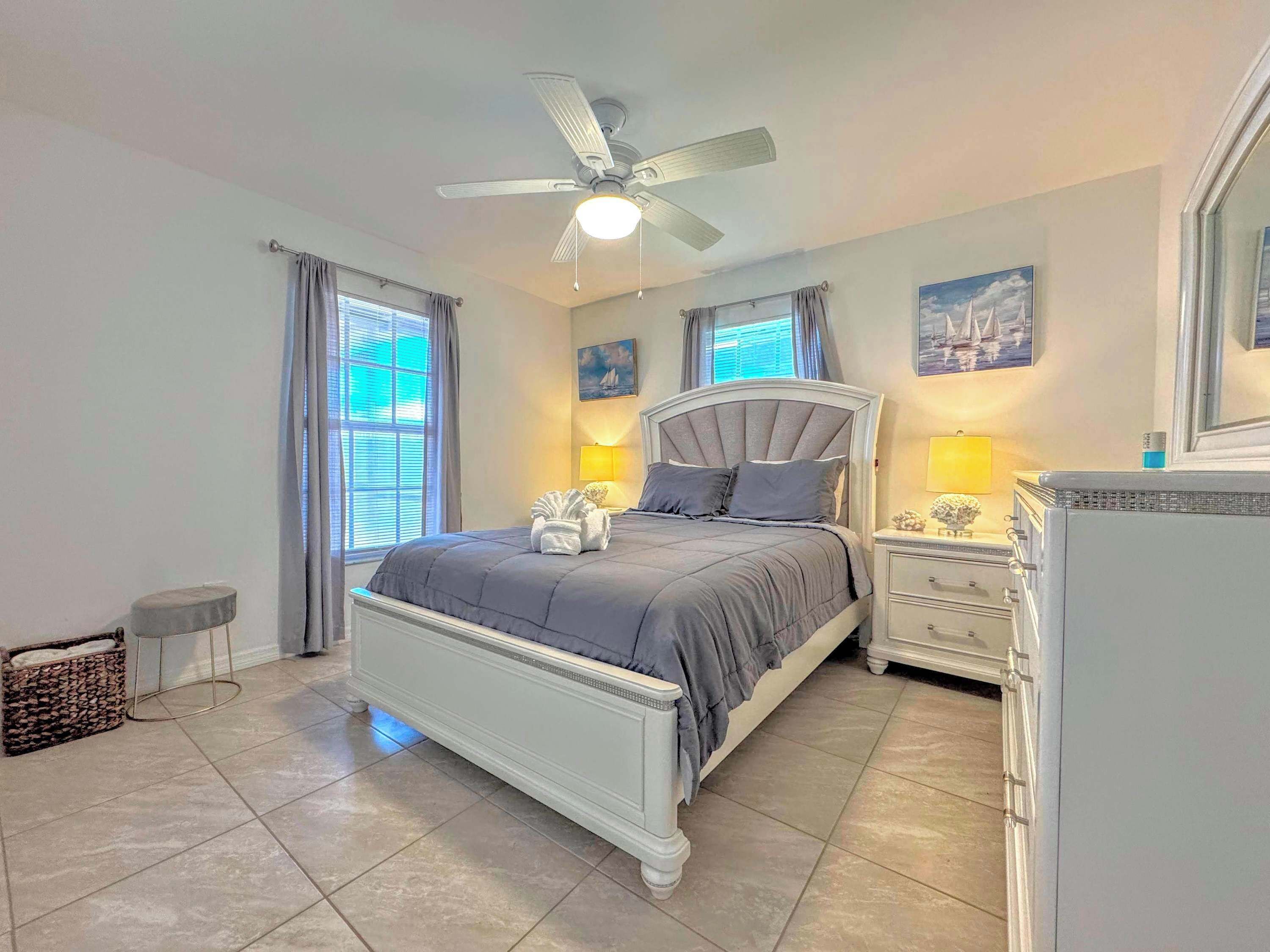 Un dormitorio con cama, mesitas de noche, ventilador de techo y ventana con cortinas.