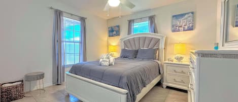 Un dormitorio con cama, mesitas de noche, ventilador de techo y ventana con cortinas.