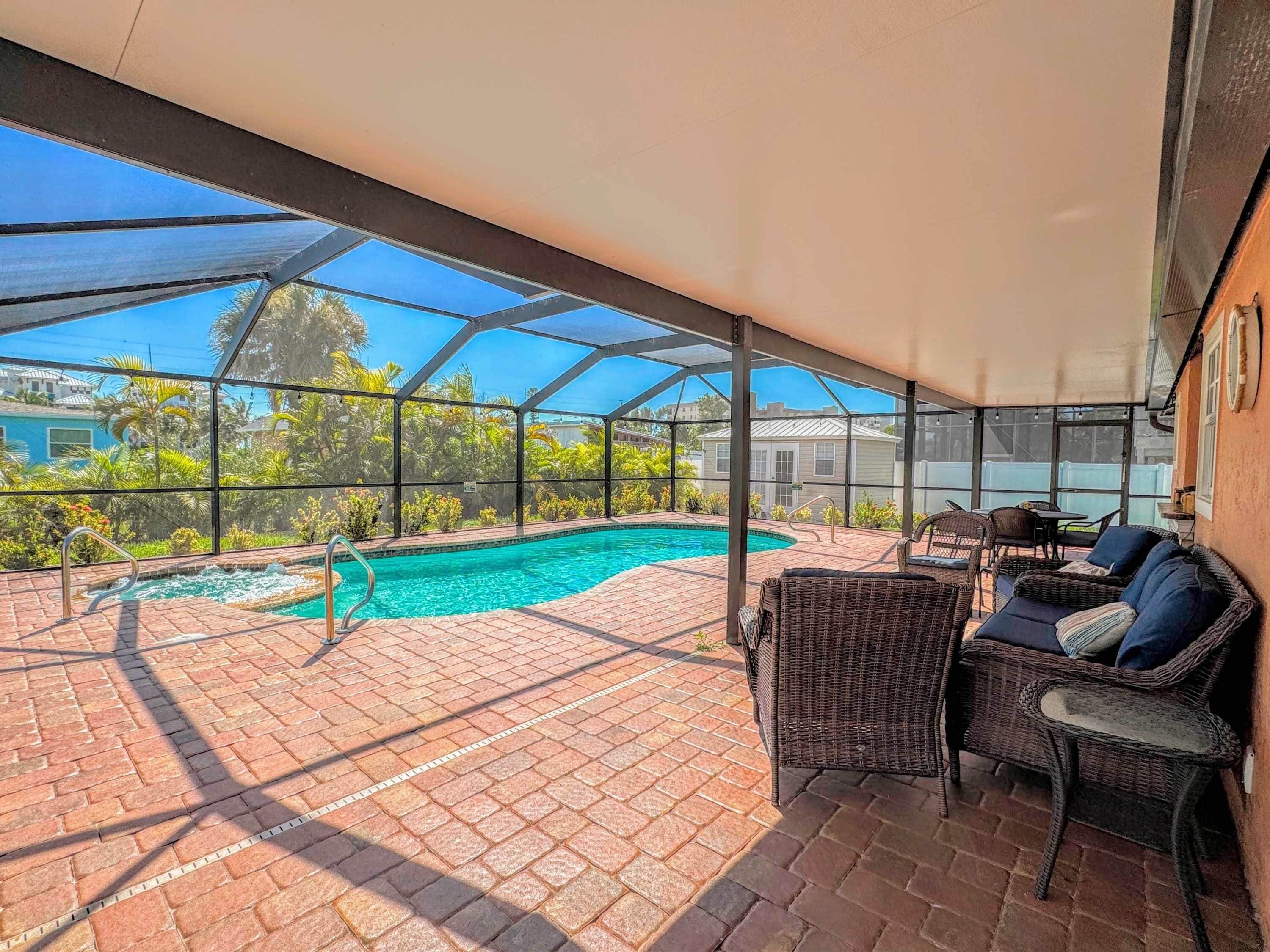 A covered outdoor swimming pool with a brick patio, wicker furniture, and a view of the surrounding area.