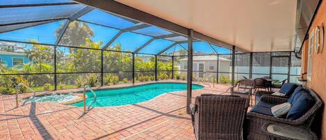 A covered outdoor swimming pool with a brick patio, wicker furniture, and a view of the surrounding area.