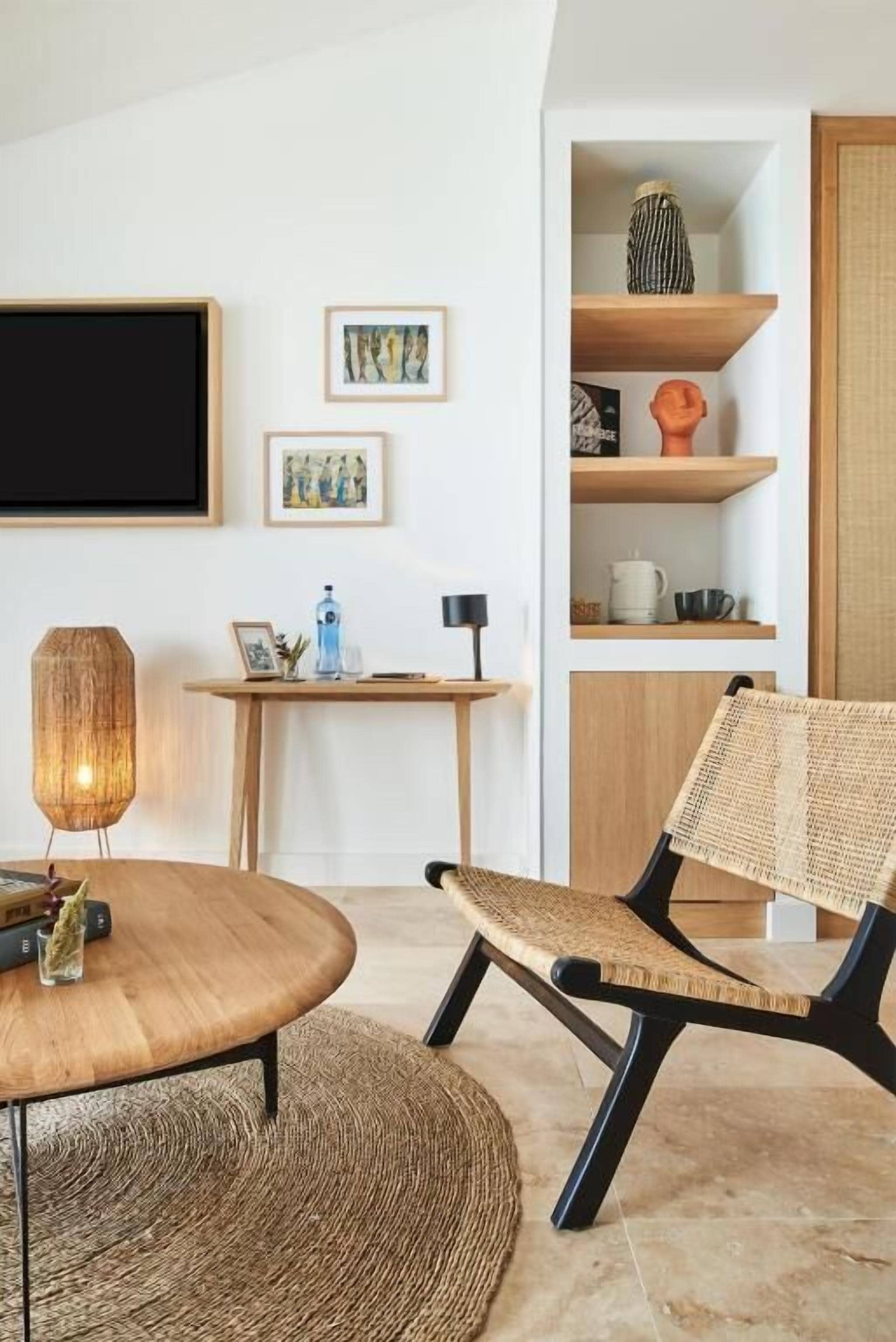 Una sala moderna con una mesa redonda de madera, una silla de fibra natural, un televisor montado en la pared y una estantería con adornos.