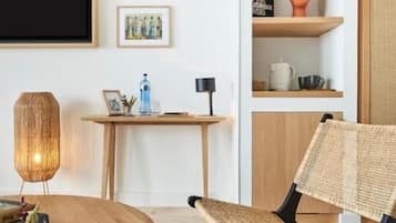 Una sala moderna con una mesa redonda de madera, una silla de fibra natural, un televisor montado en la pared y una estantería con adornos.