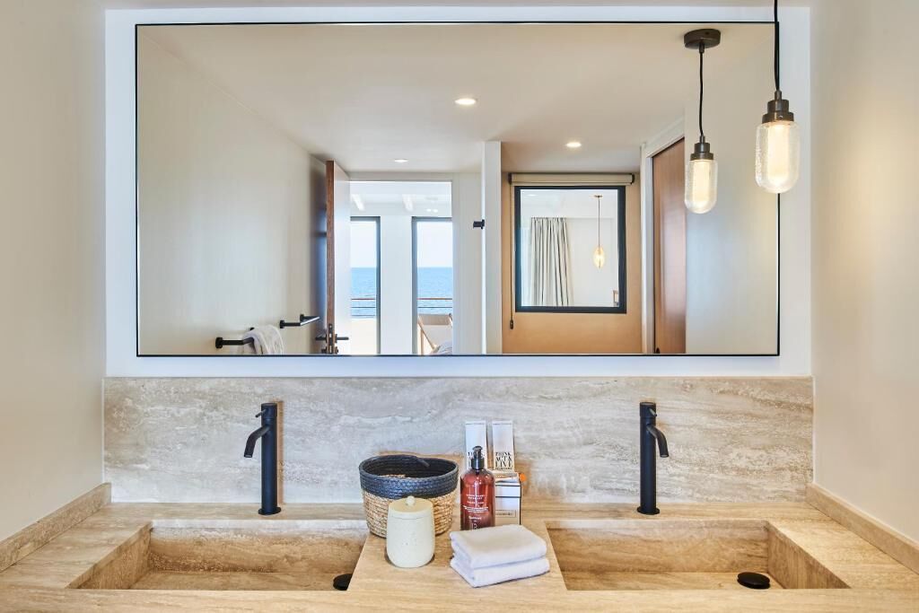 A bathroom with a large mirror, two sinks, and a view of the sea through an open door.