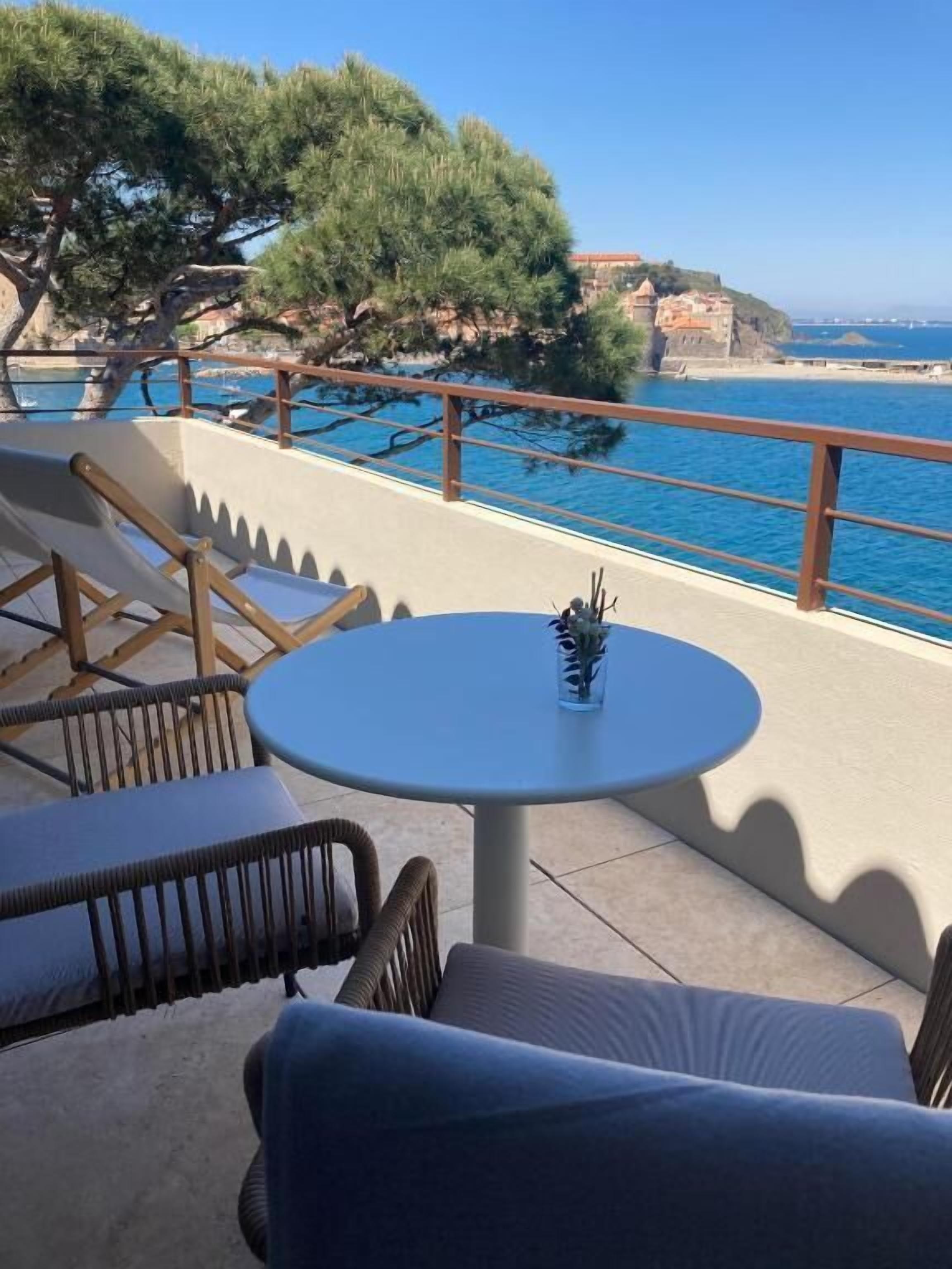 A terrace with wicker furniture and a table, overlooking a coastal area with trees and cliffs.