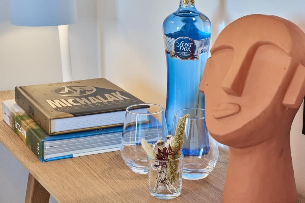 Ett träbord med en bok som har titeln ”MICHAELAK”, en blå flaska med etiketten ”FONT DOR MAXIMUM”, två genomskinliga glas och en terrakottabyst.