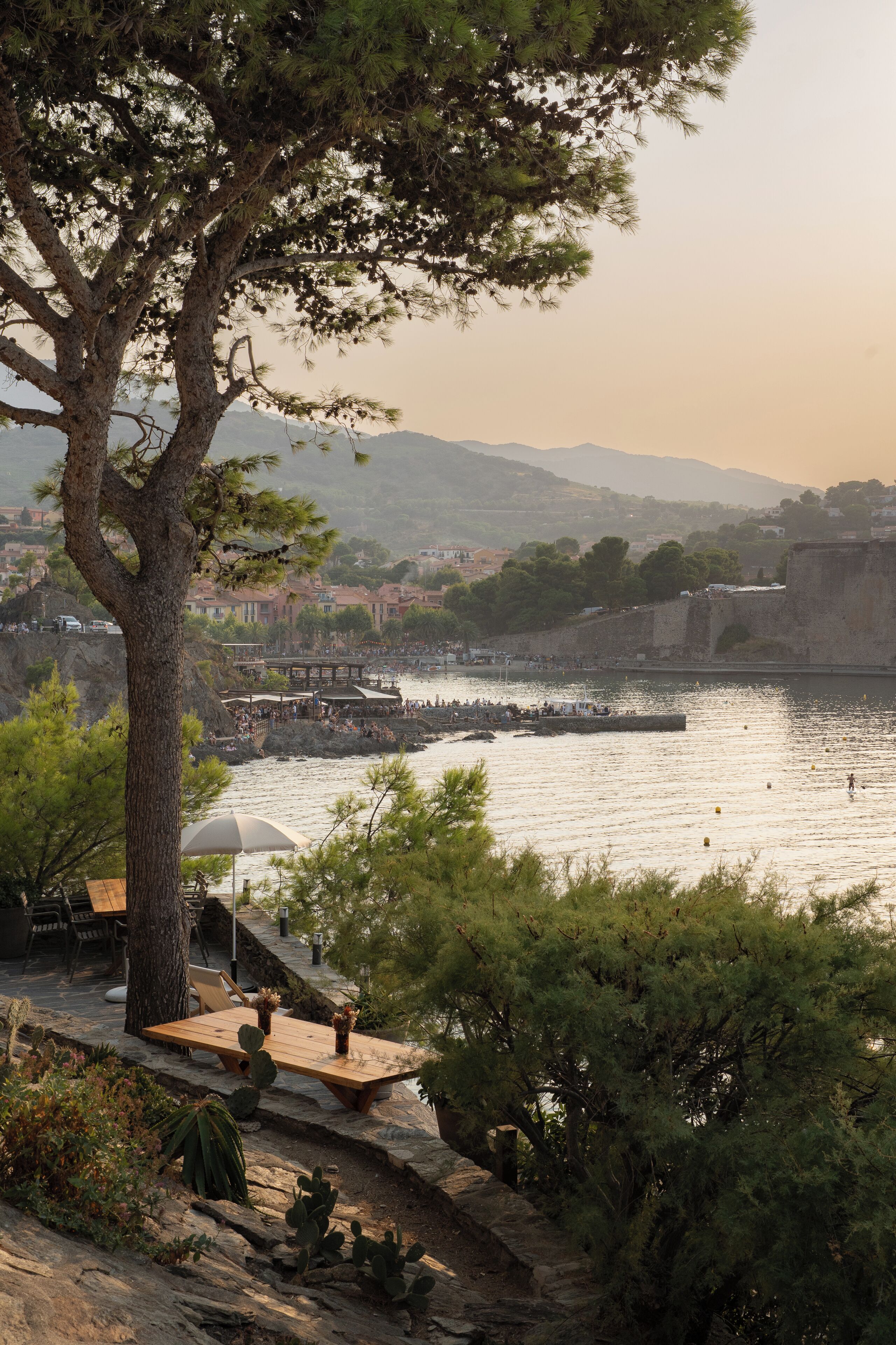 A coastal town with a stone table and chairs under a tree, overlooking a calm sea and a historic fort.