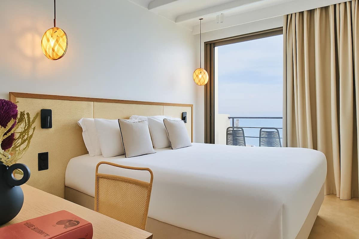 Un cuarto de hotel con una cama grande, una zona de comedor con mesa y sillas, y un balcón con vista al mar.