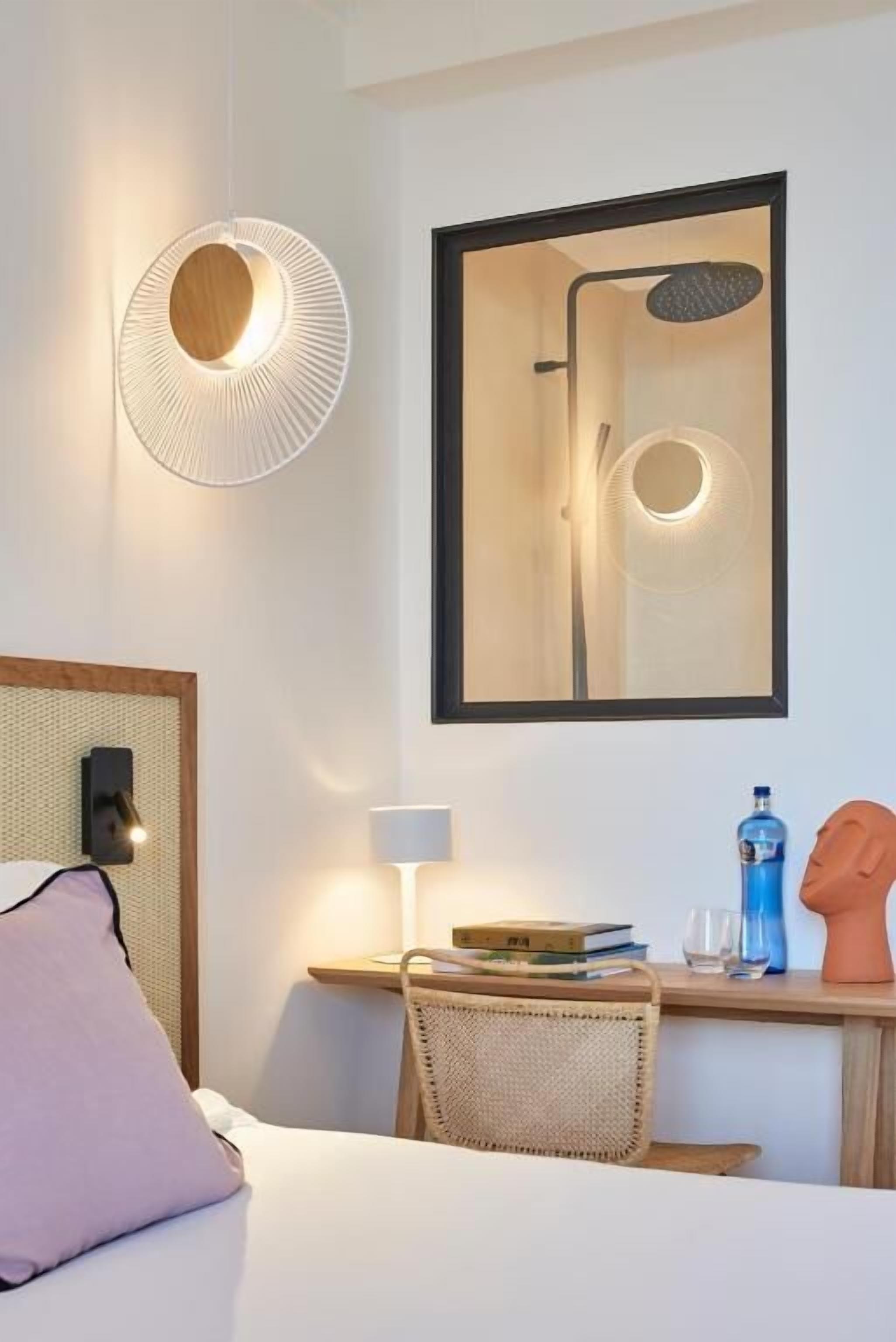 Una habitación de hotel moderna con cama, escritorio con lámpara, una silla, un cuadro enmarcado de una ducha y una lámpara de pared.