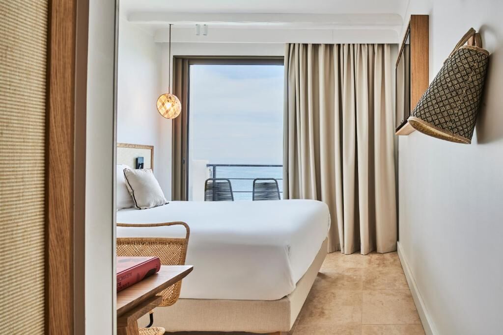 A hotel room with a bed, a small table with a book, a chair, a balcony with curtains, and a hanging lamp.