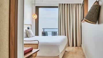 A hotel room with a bed, a small table with a book, a chair, a balcony with curtains, and a hanging lamp.