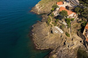 Vue aérienne d’une propriété située sur la côte, avec un rivage rocheux, un chemin pavé et un bâtiment surmont é d’un toit recouvert de tuiles rouges.