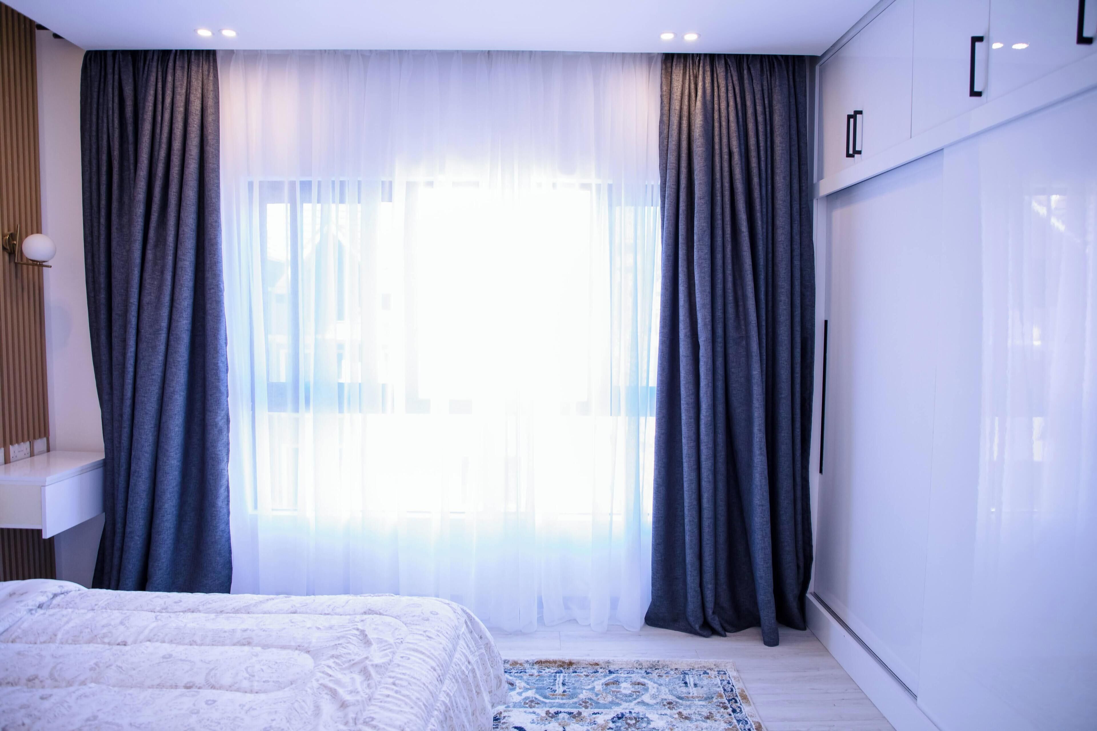 A bedroom with a bed, a bedside table, a wardrobe, and a window with sheer curtains.