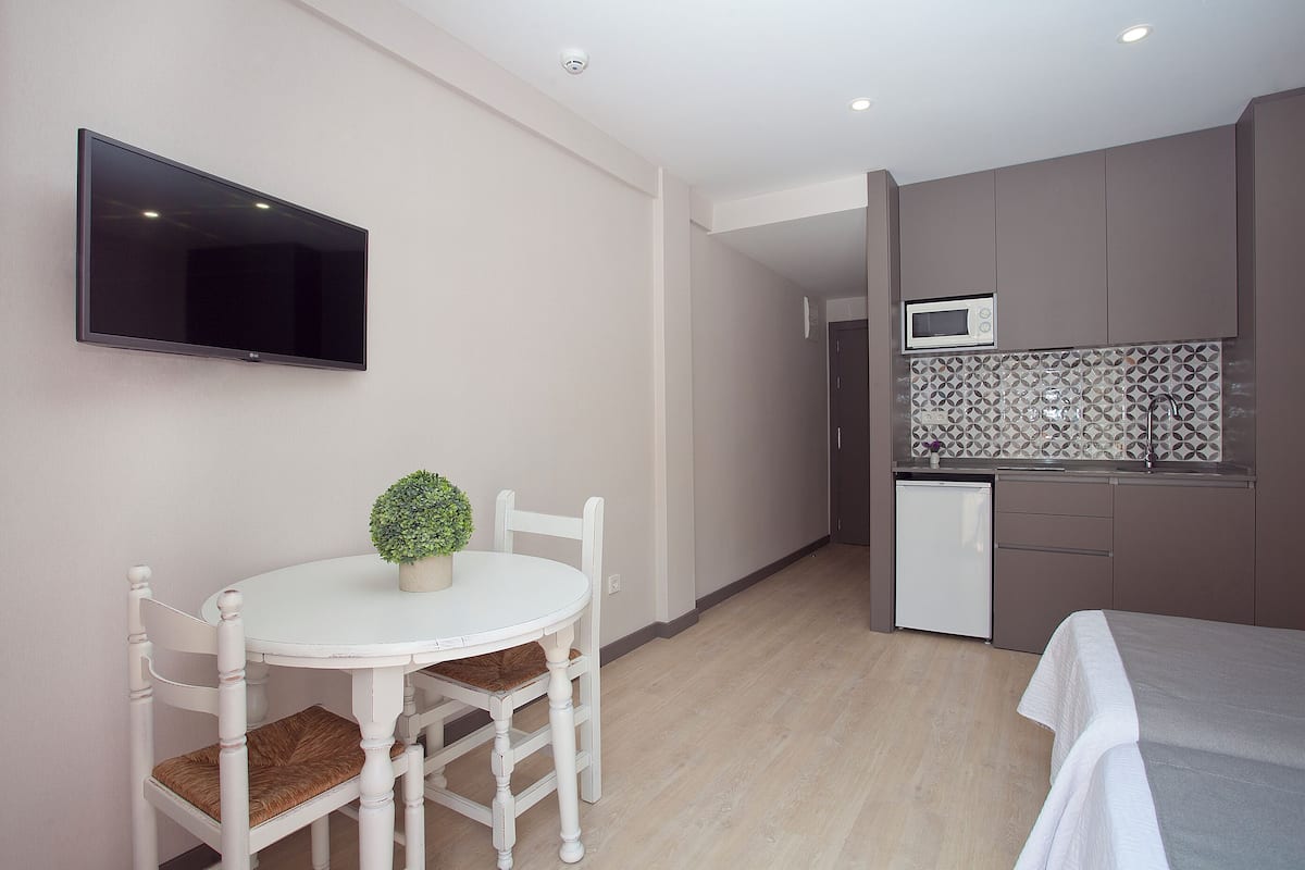 A compact room with a dining table, chairs, a small potted plant, a flat-screen TV mounted on the wall, a kitchenette with a microwave, and a bed with a white bedspread.