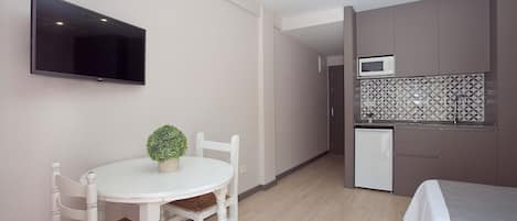 A compact room with a dining table, chairs, a small potted plant, a flat-screen TV mounted on the wall, a kitchenette with a microwave, and a bed with a white bedspread.