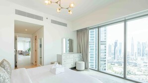 A modern hotel room with a large bed, a dresser, and a view of the cityscape through floor-to-ceiling windows.