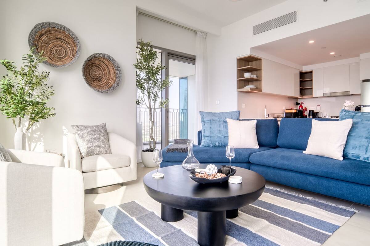 A modern living room with a blue sofa, a white armchair, and a coffee table. There is a kitchen area visible in the background.