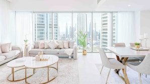 A modern living room with a sofa, coffee table, and dining table, large windows with a city view, and minimal decor.