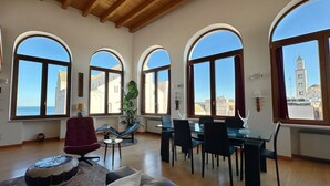 A spacious living area with a high wooden ceiling, large arched windows, a dining table with chairs, and a view of a cityscape with a tower. - Dimora Riccardi San Nicola (Bari)
