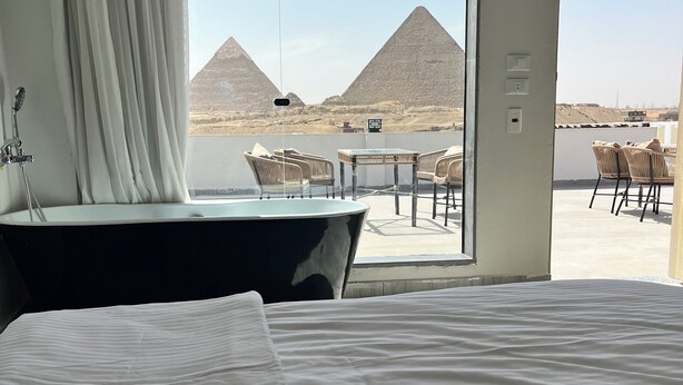 A hotel room with a large bed, a bathroom with a bathtub, and a view of the pyramids.
