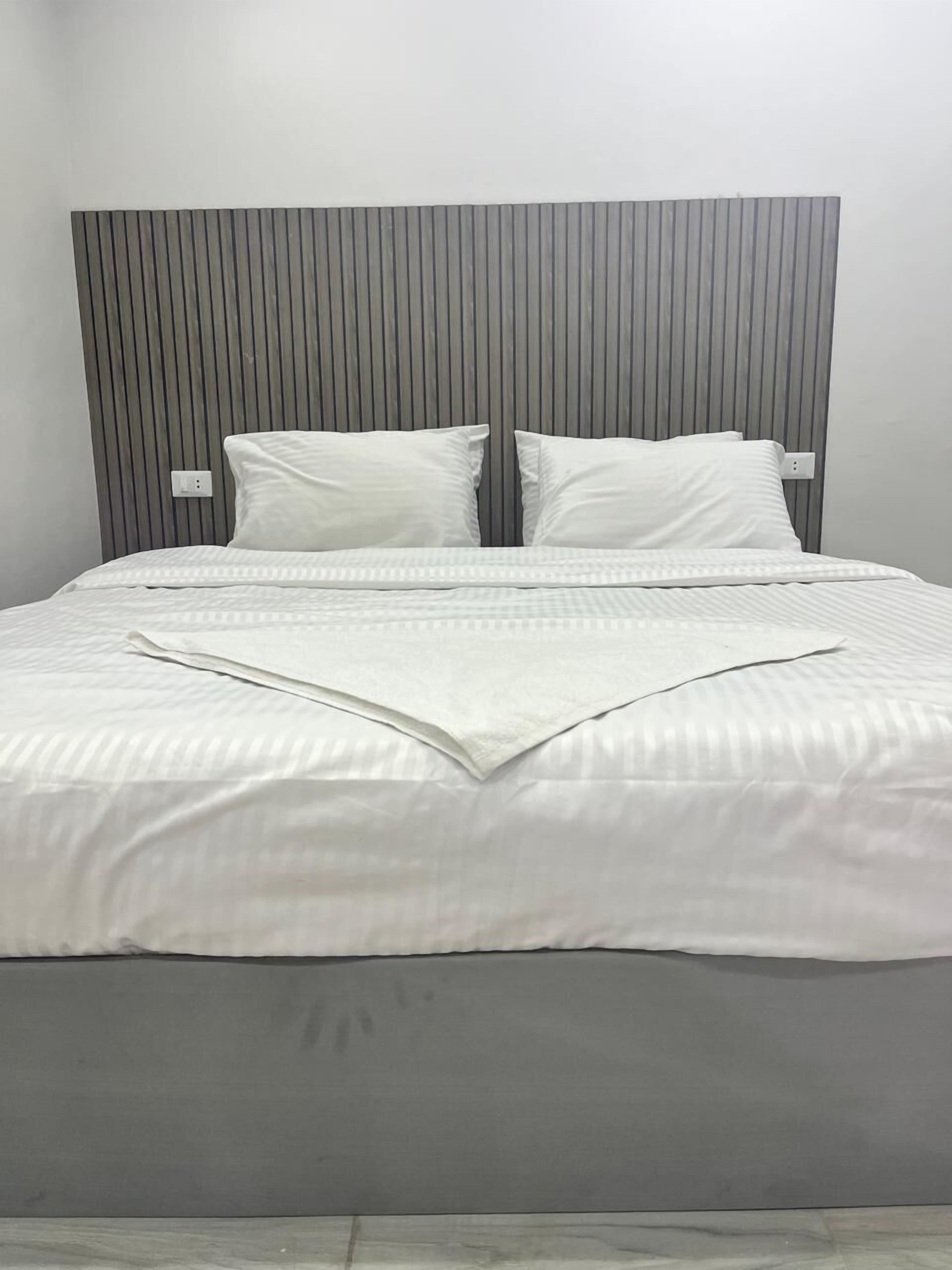 Een net opgemaakt bed met witte lakens en kussens, een grijs hoofdbord en een effen muur.
