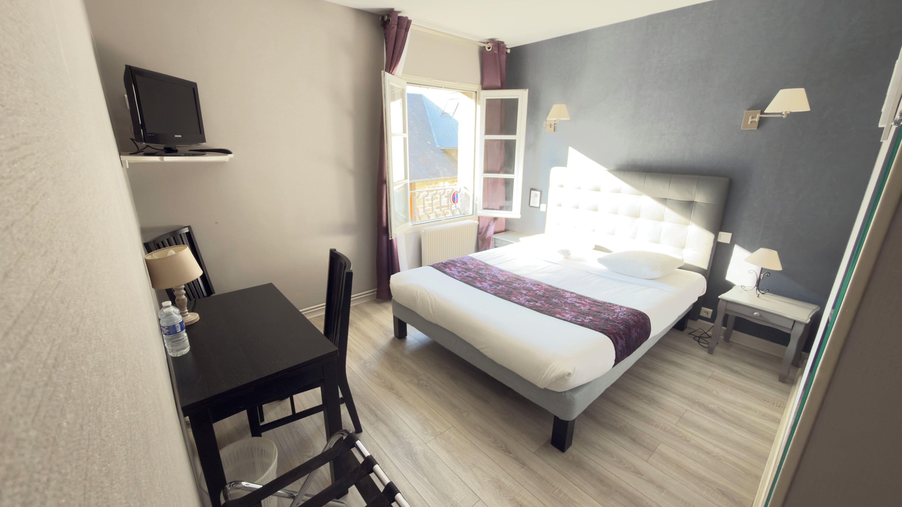 A hotel room with a bed, bedside tables, a desk with a chair, a television, and a window with curtains.