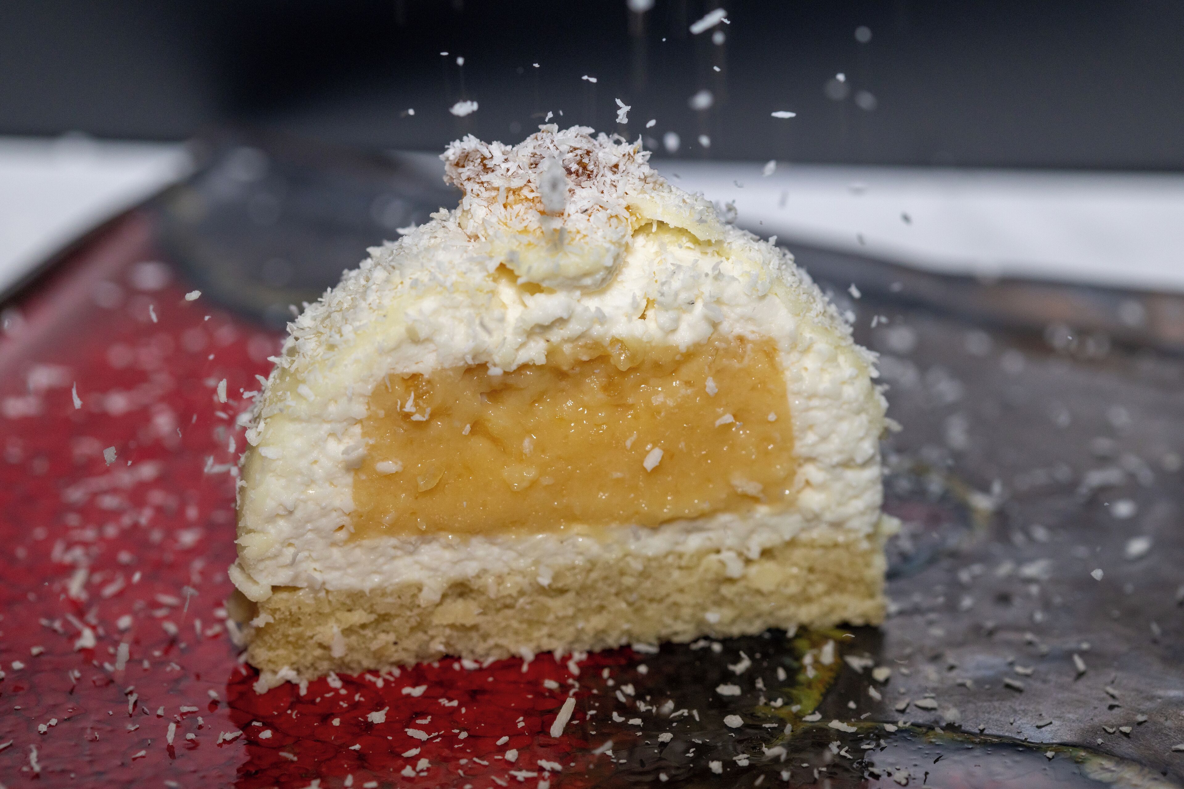 A dessert with a creamy topping and coconut flakes, placed on a red surface with a blurred background.