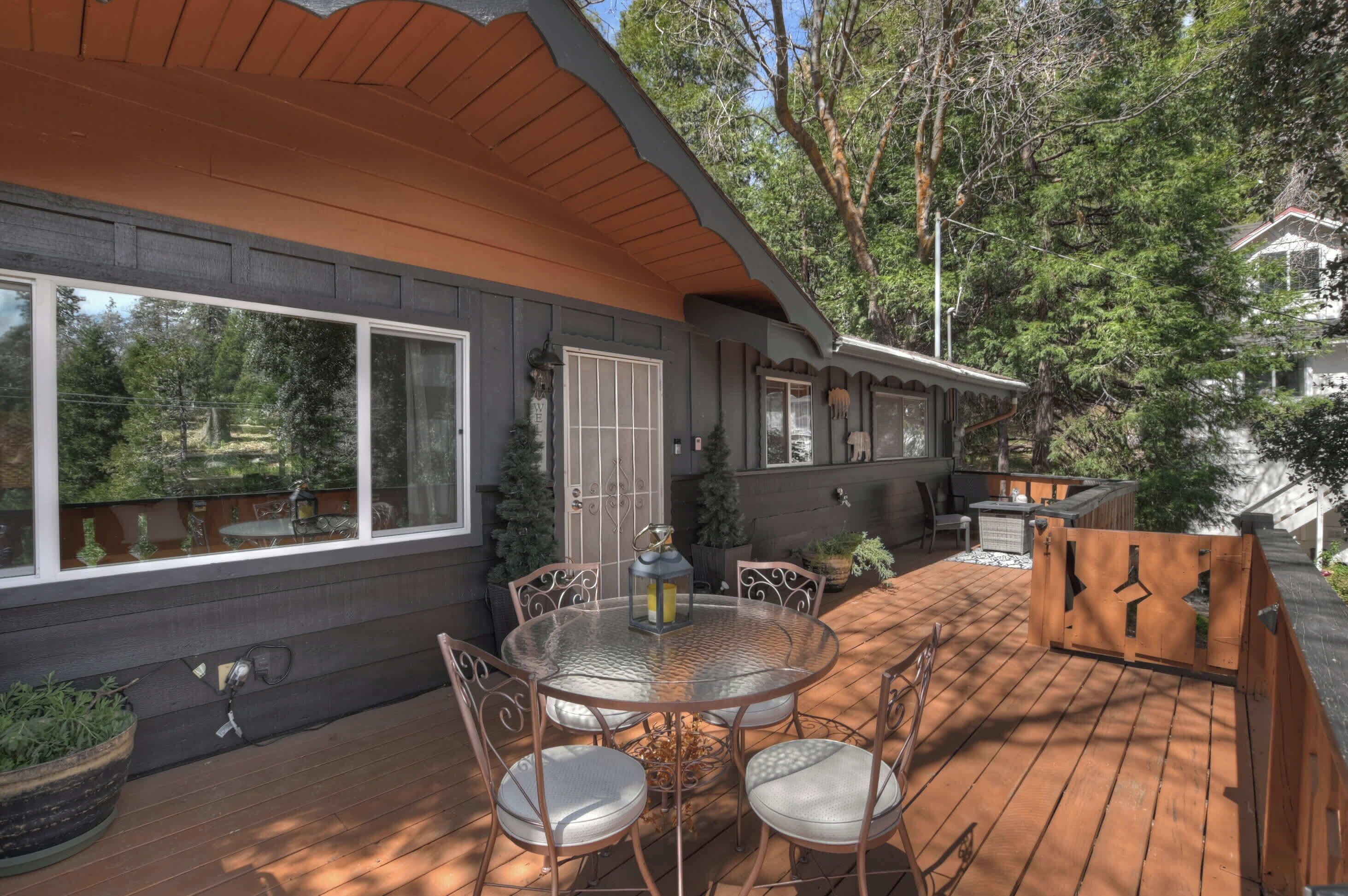 A wooden deck with a round glass table and chairs, surrounded by potted plants and a small outdoor kitchen area.