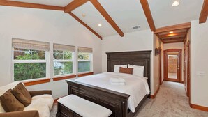 A bedroom with a large bed, wooden beams on the ceiling, a window with a view of greenery, a bench, and a door leading to another room.